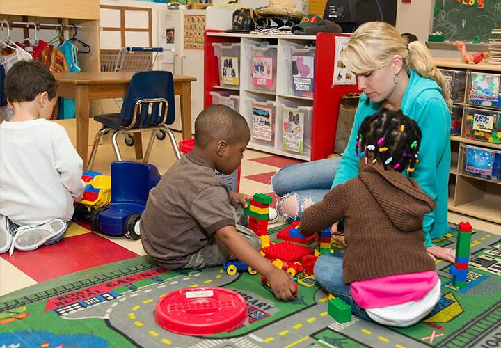 A teacher sits with little children playing with large LEGO DUPLO bricks in a daycare.