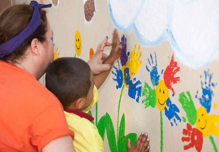 A preschool teacher helps a little boy paint flowers on a colorful exterior mural using his handprint.